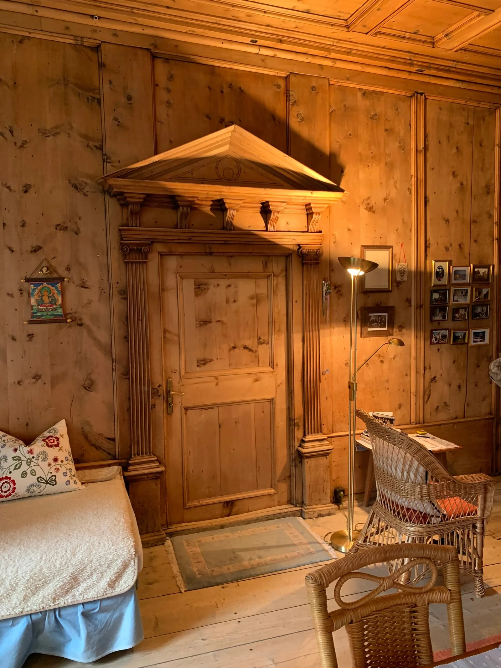 Holzvertäfelte Wand mit Tür, Säulen und einem Giebelportal darüber, davor ein Bett, ein Korbstuhl und eine Stehlampe