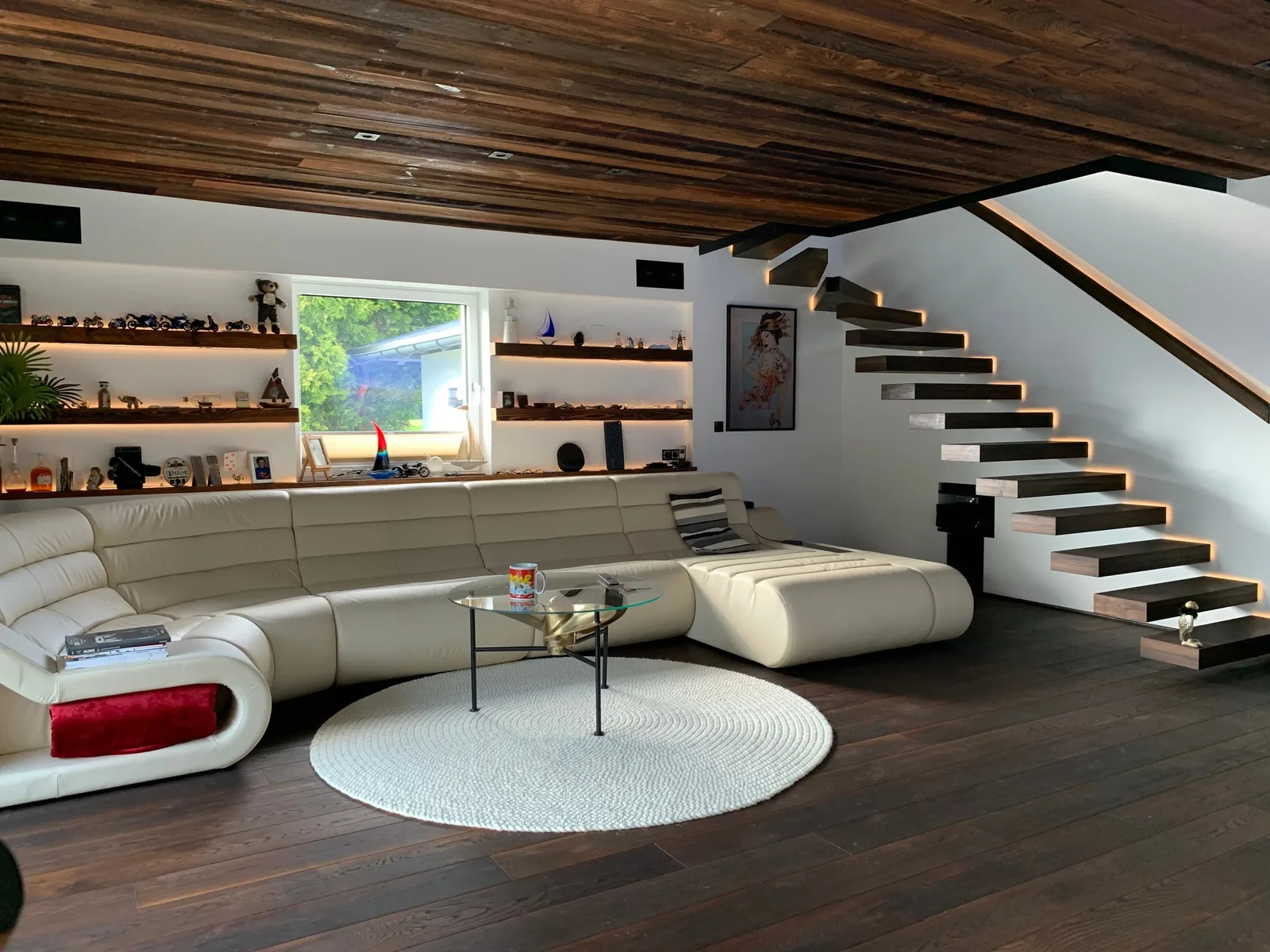 Wohnraum mit dunkler Holzdecke, beleuchteten Wandborden und schwebender Treppe