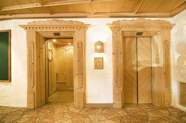 Zwei Lifttüren, jede mit einem geschnitzten Holzportal mit Ornamenten, dazwischen eine Wandleuchte