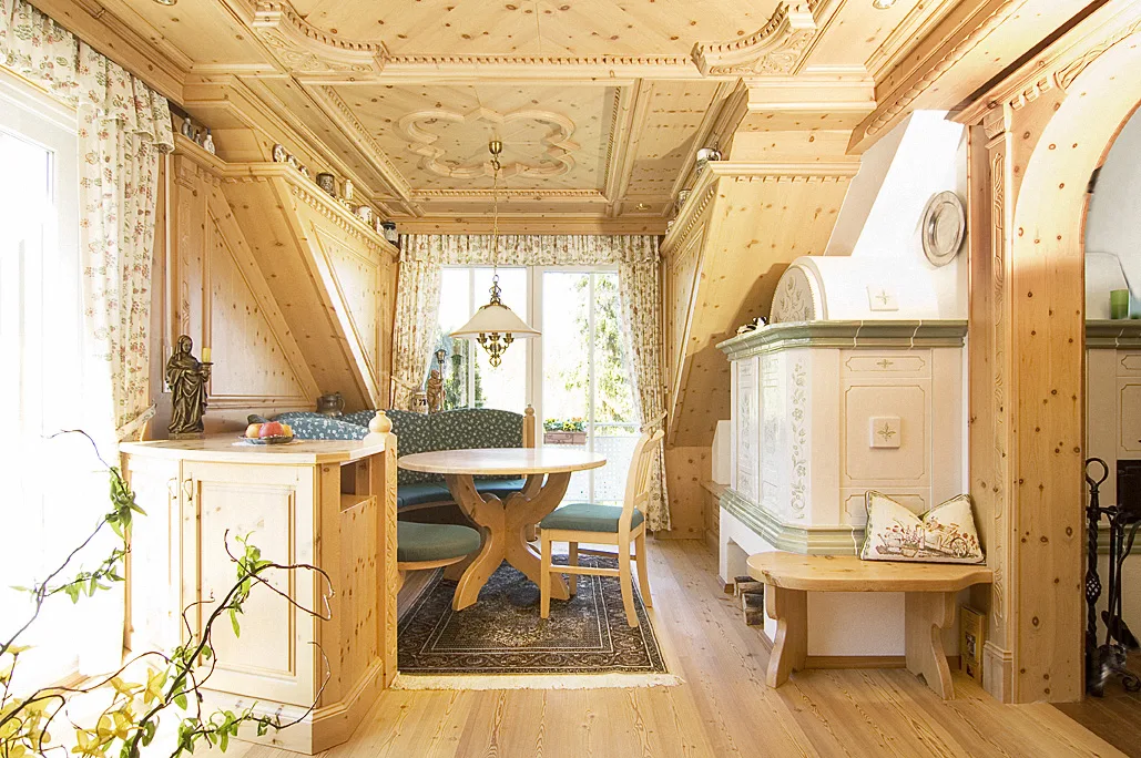 Stube aus hellem Holz mit geschnitzter Kassettendecke, Eckbank, rundem Tisch und Kachelofen