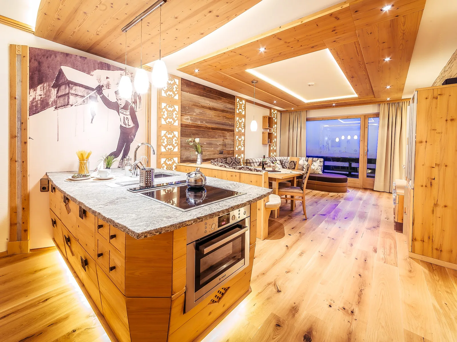 Kücheninsel aus Holz mit heller Steinplatte, dahinter geschnitzte Paneele, Essplatz und eine Holzdecke mit Lichtband